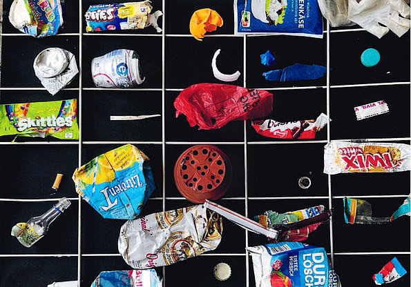 Verschiedene Müll- und Verpackungsreste wie zerknüllte Snack- und Getränkepackungen, Flaschendeckel, Kaugummiverpackungen und Zigarettenstummel liegen in einem Gitterraster auf schwarzem Hintergrund.