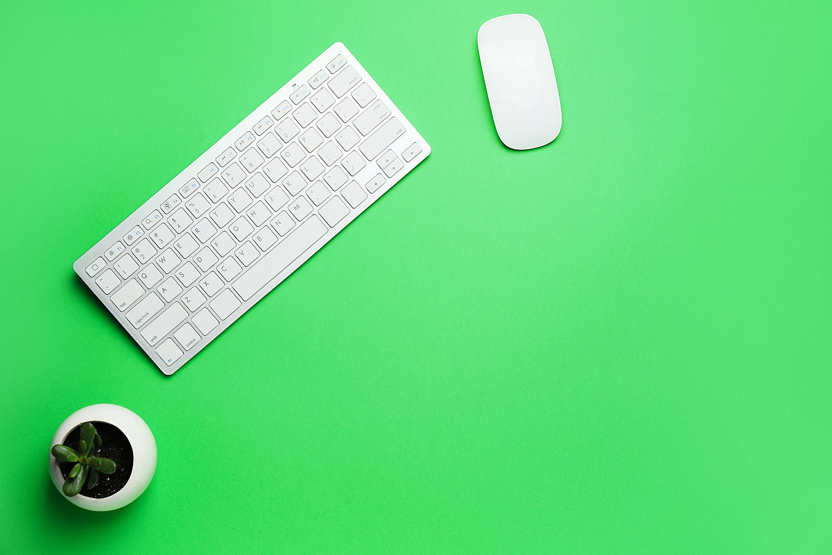 Weiße Computertastatur, Maus und kleine Pflanze auf grünem Schreibtisch – symbolisch für digitale Bildungsangebote für Erwachsene.