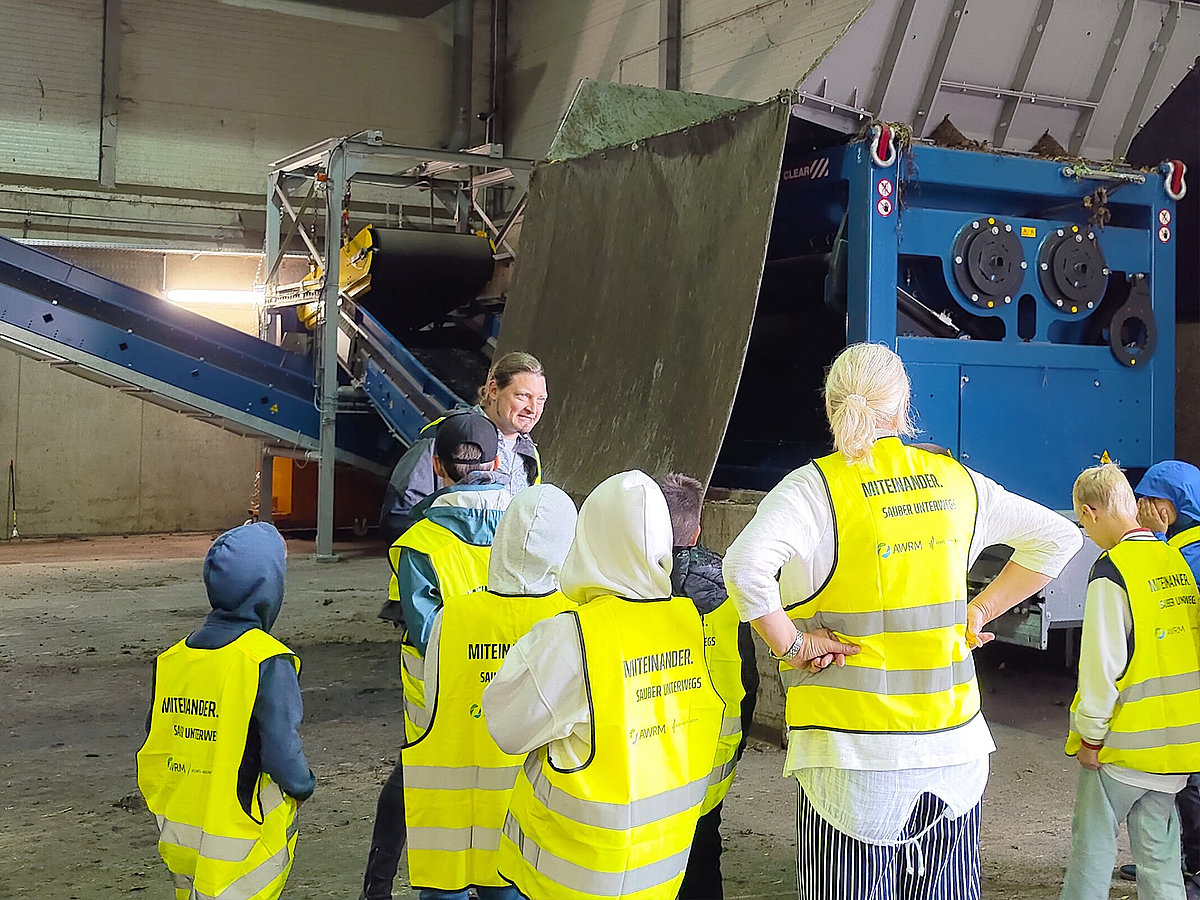 Gruppe von Kindern mit gelben Warnwesten bei einem Erlebnistag in einer Recyclinganlage; ein Mitarbeiter erklärt eine große Maschine zur Abfallverarbeitung.