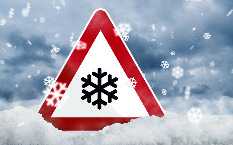 Warnschild mit Schneeflocken-Symbol steckt in einer verschneiten Landschaft, während dichter Schneefall vor einem grau bewölkten Himmel niedergeht.
