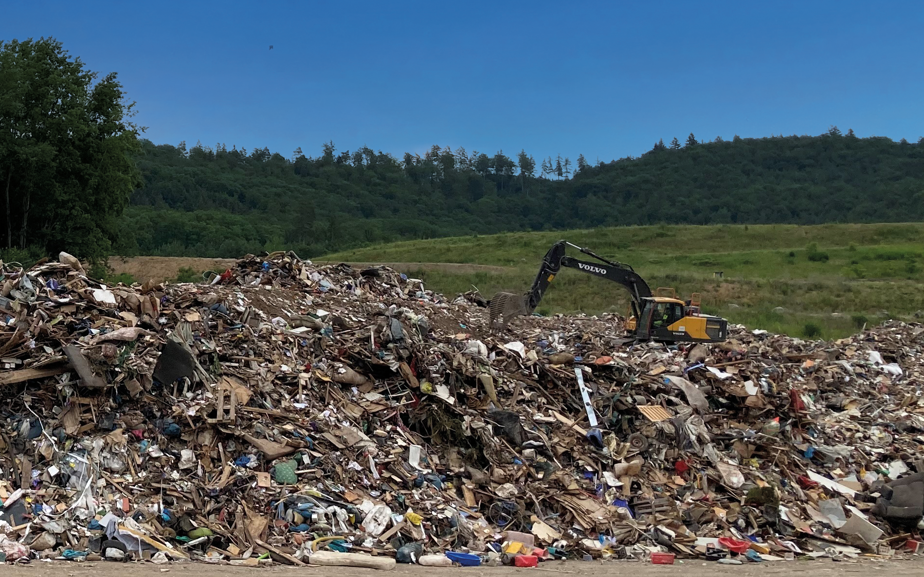 Müllberg mit Bagger vor blauem Himmel, im Vordergrund Asphalt