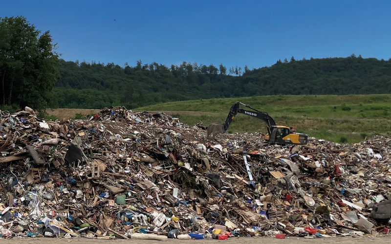 Müllberg mit Bagger vor blauem Himmel, im Vordergrund Asphalt
