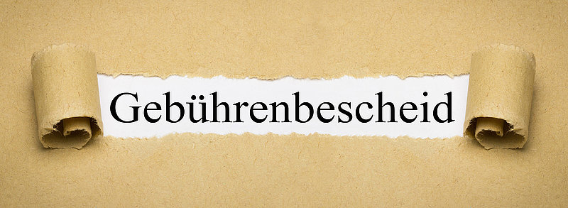 Aufgerissenes Packpapier legt das Wort „Gebührenbescheid“ in schwarzer Serifenschrift frei.