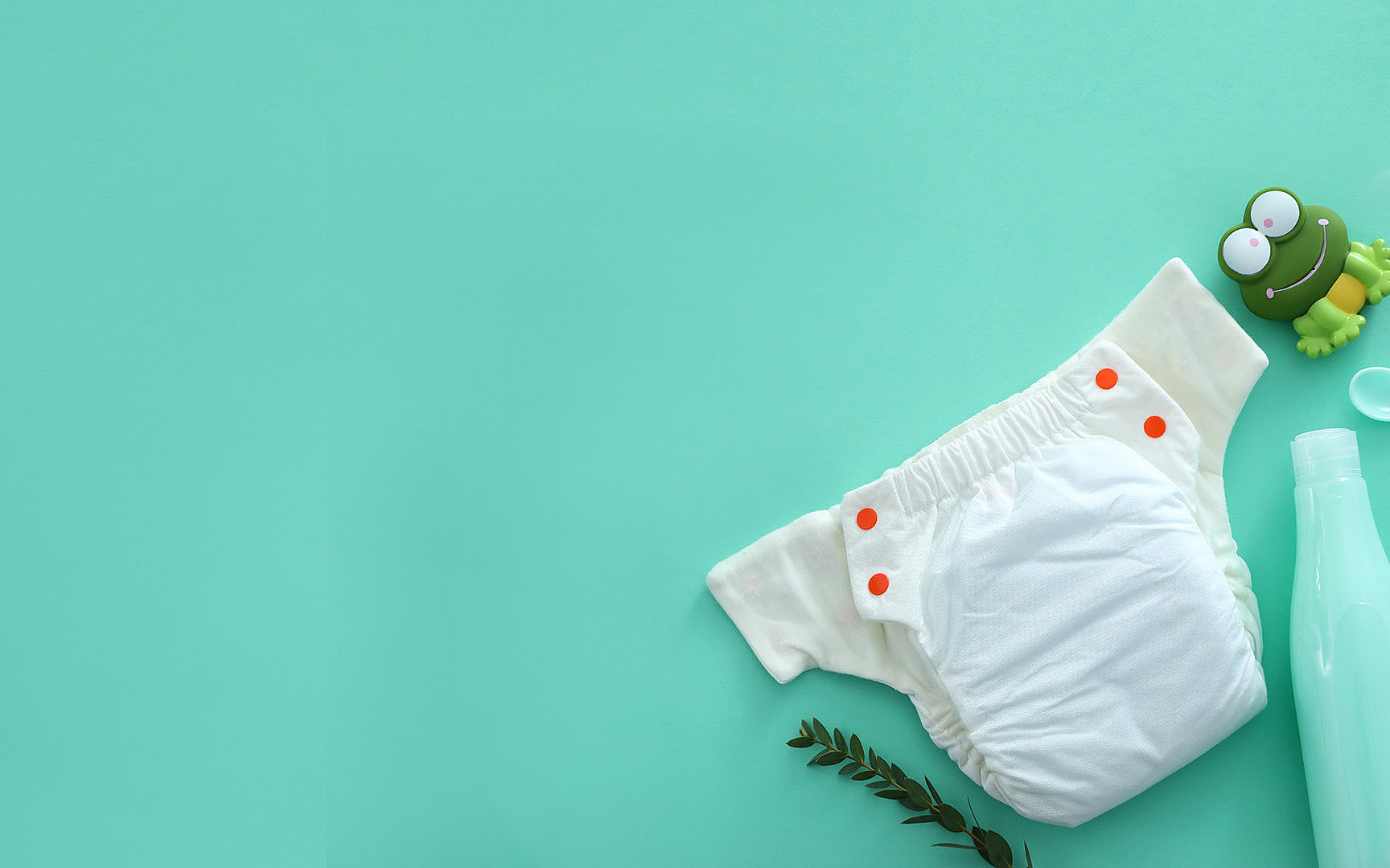 Weiße Stoffwindel mit orangefarbenen Druckknöpfen auf mintgrünem Hintergrund, daneben eine grüne Quietschefroschfigur und eine Babyflasche