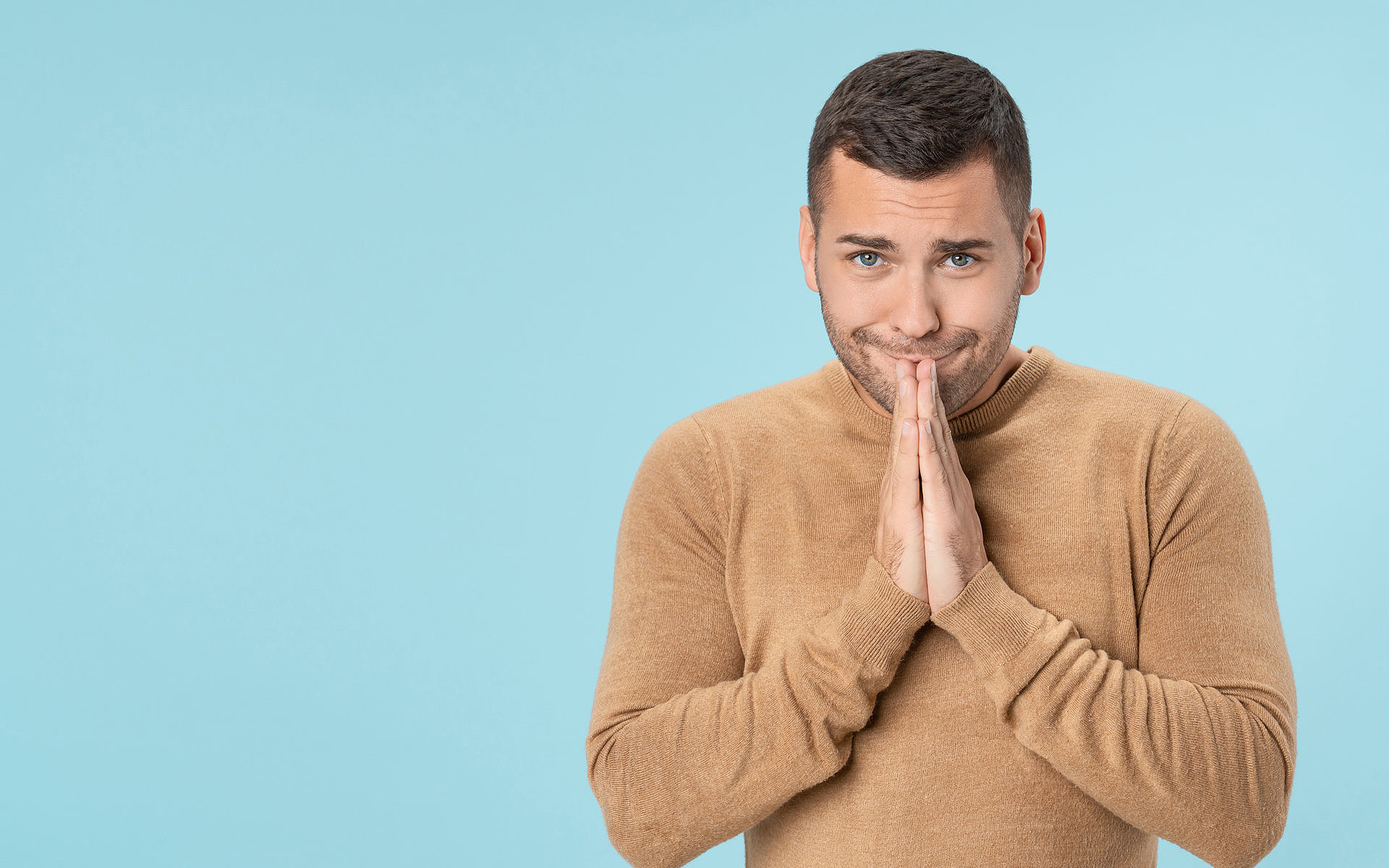 Junger Mann mit braunem Pullover vor hellblauem Hintergrund, der die Hände vor dem Gesicht faltet und entschuldigend schaut.