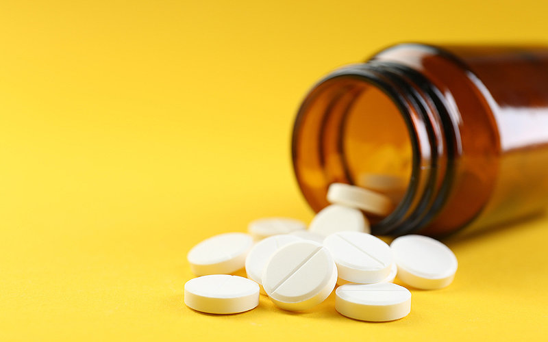 Eine braune Glasflasche liegt geöffnet vor einem gelben Hintergrund, vor der Flaschenöffnung liegen Tabletten