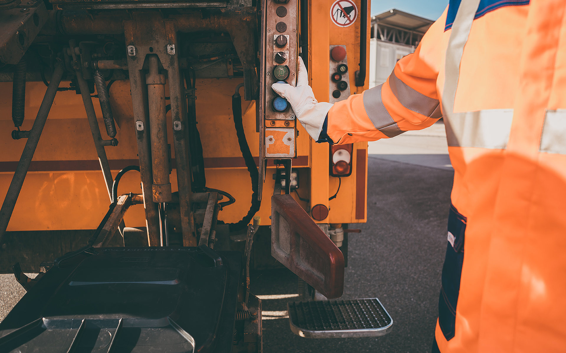 Mitarbeiter der Müllabfuhr in orangefarbenem Arbeitsanzug bedient Steuerung am Müllwagen, bereit zur Abholung der Mülltonne.