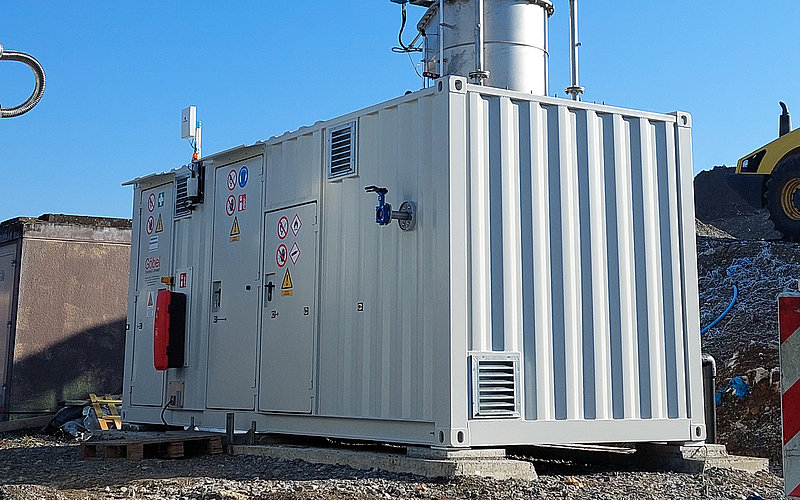 Technische Anlage in Containerbauweise mit hohen Edelstahlrohren zur Schwachgasverbrennung. Die Anlage steht auf einem geschotterten Platz unter blauem Himmel.