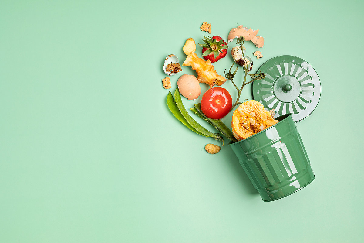 Ein umgekippter grüner Bioabfalleimer auf grünem Untergrund, aus dem organische Küchenabfälle wie Obst- und Gemüsereste sowie Eierschalen herausgefallen sind.