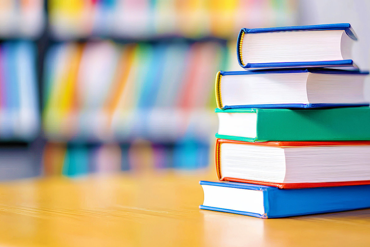Gestapelter Bücherstapel mit bunten Einbänden auf einem Tisch, im Hintergrund unscharfe Bücherregale.