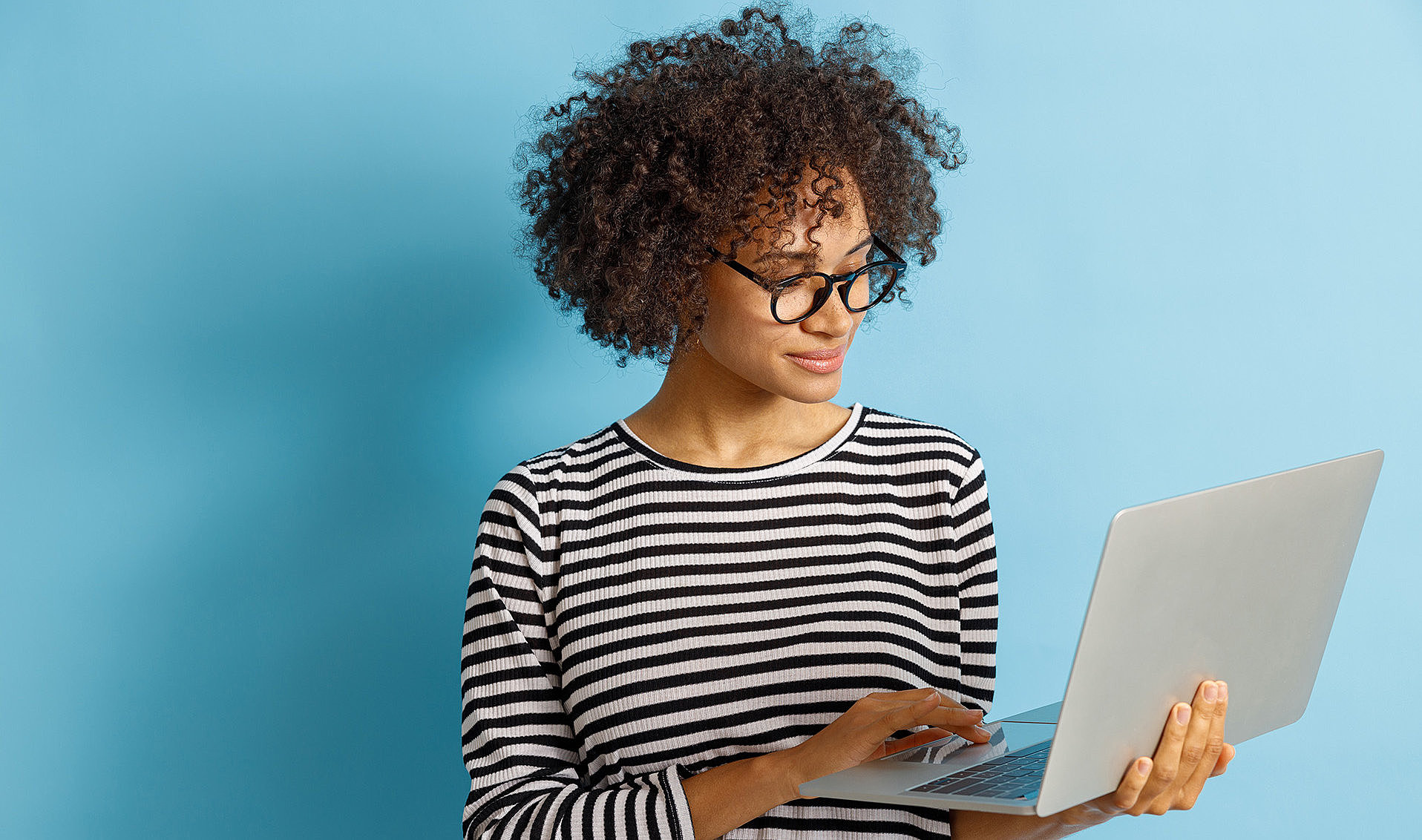 Junge Frau mit Locken und Brille hält einen Laptop in der Hand und tippt darauf vor hellem Hintergrund.