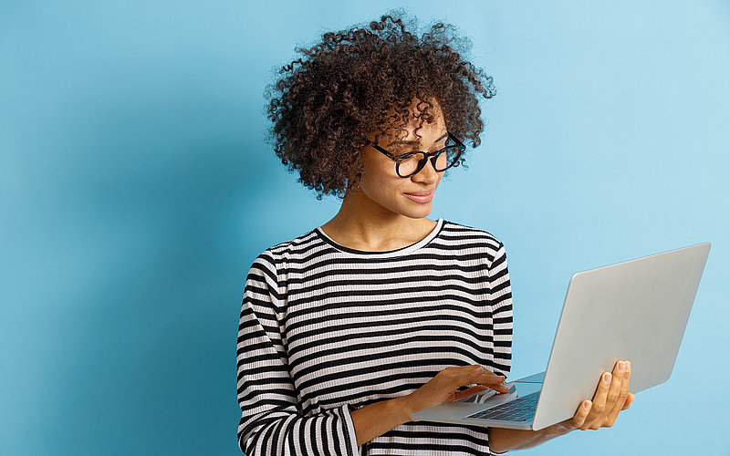 Junge Frau mit Locken und Brille hält einen Laptop in der Hand und tippt darauf vor hellem Hintergrund.