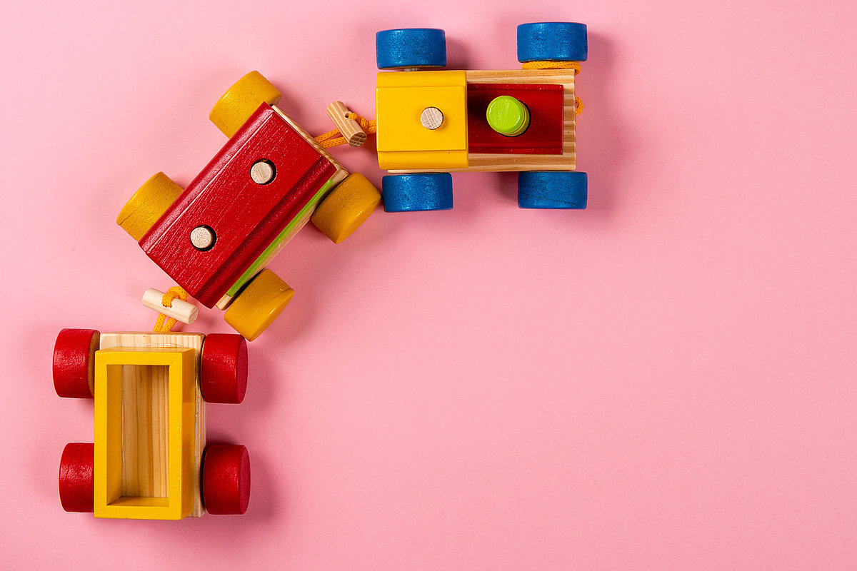 Drei bunte miteinander verbundene Holzspielzeugautos mit roten, gelben, blauen und grünen Elementen stehen auf einem rosa Hintergrund.