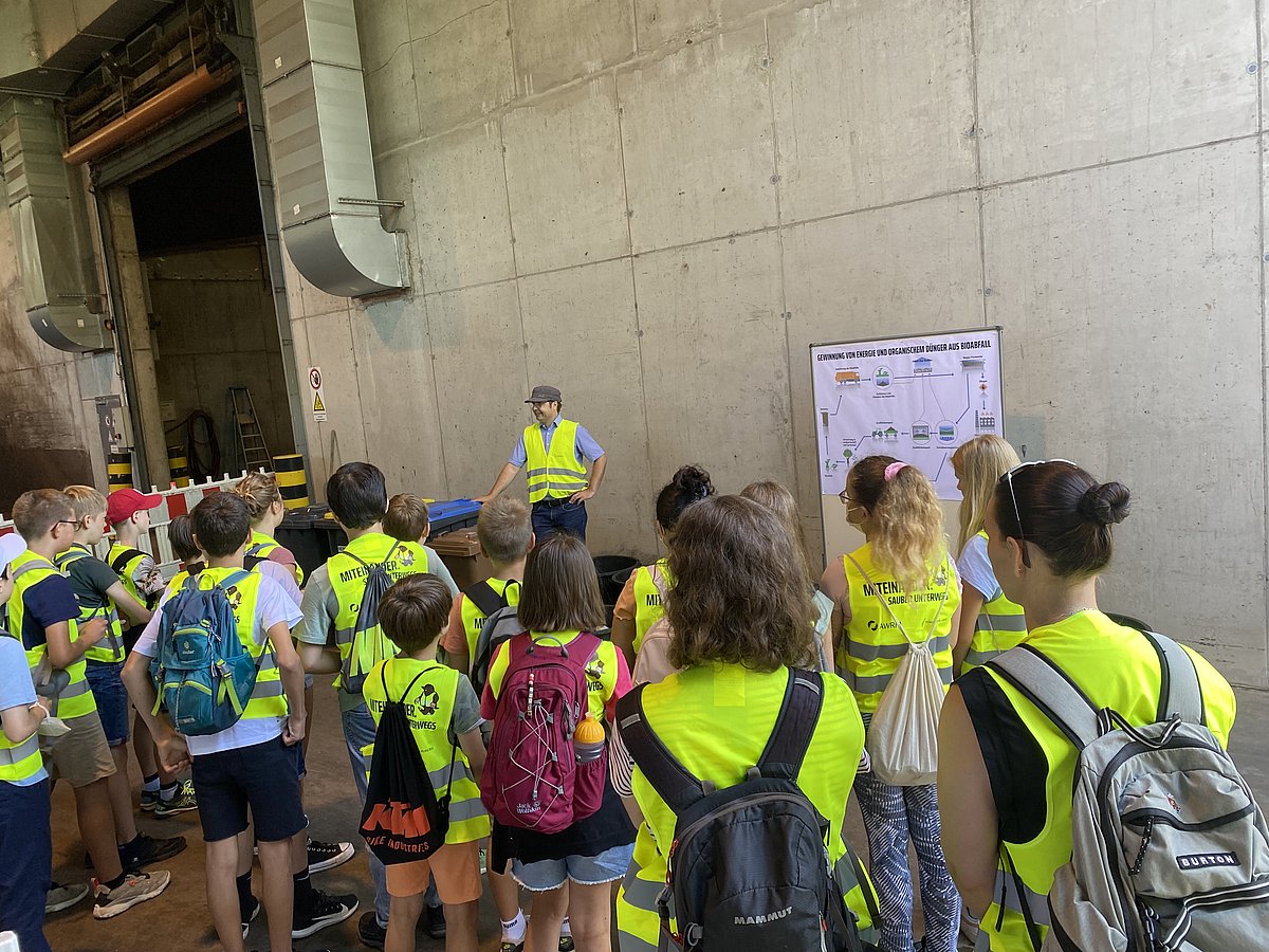Ein Sachbearbeiter der Schulberatung der AWRM erklärt einer Gruppe von Kindern in Warnwesten bei einer Führung ein Schaubild zur Energie- und Düngergewinnung aus Bioabfall.