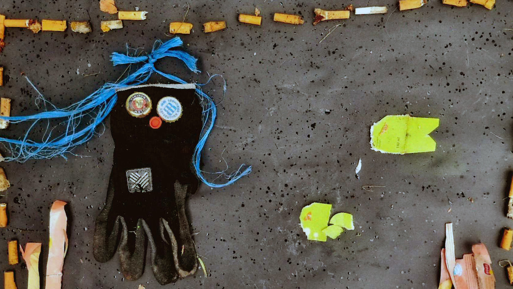 Aus Müll gestaltetes Bild: eine schwarze Handschuhfigur mit blauen Fäden als Haare, umrahmt von Zigarettenstummeln und kleinteiligem Abfall.