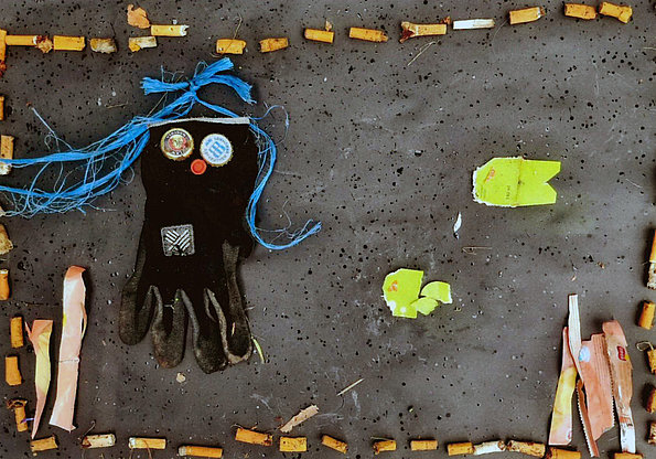 Aus Müll gestaltetes Bild: eine schwarze Handschuhfigur mit blauen Fäden als Haare, umrahmt von Zigarettenstummeln und kleinteiligem Abfall.