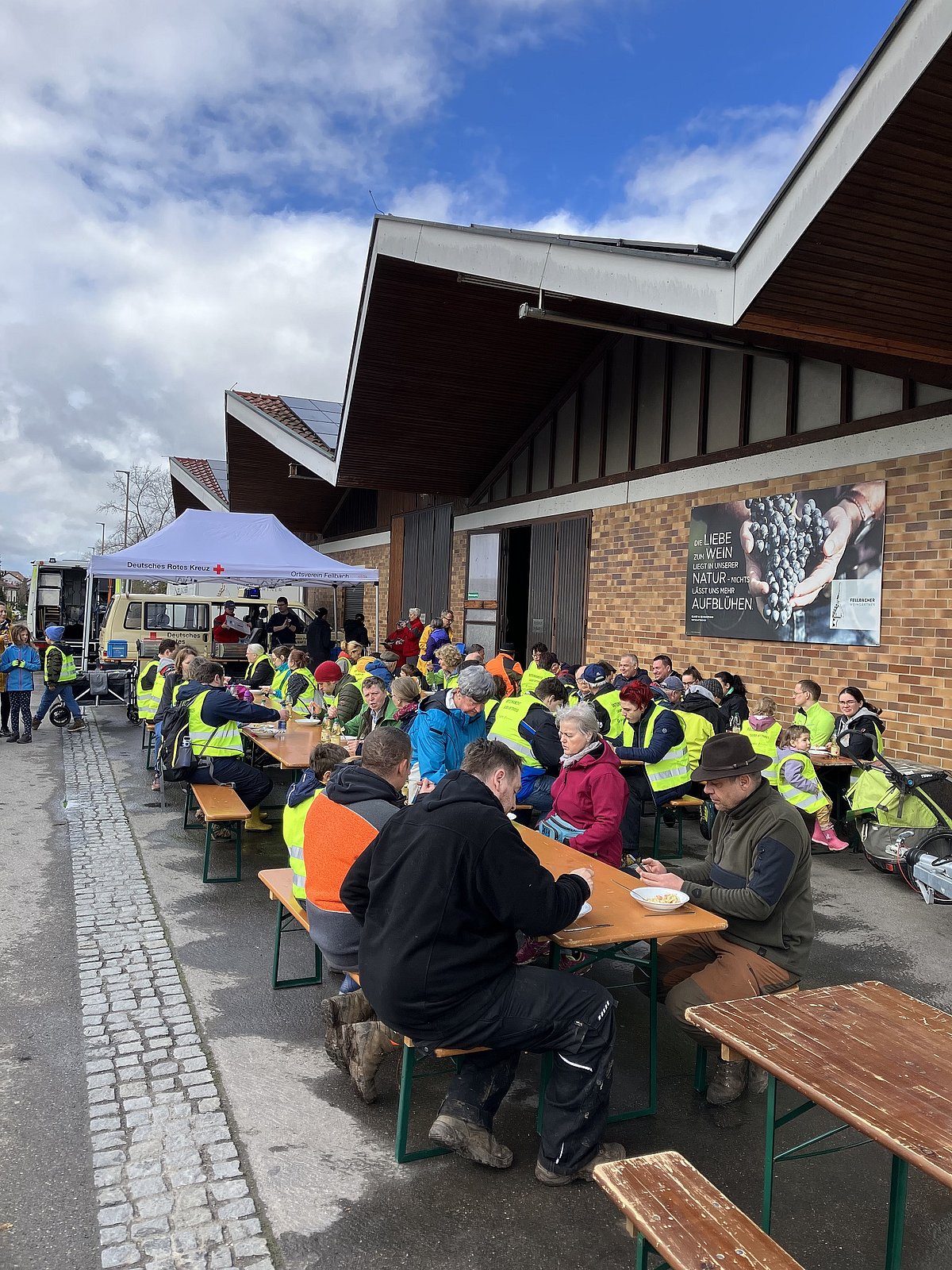 Teilnehmende der Kreisputzete in Fellbach sitzen in Warnwesten an Biertischen vor einer Halle und essen gemeinsam.