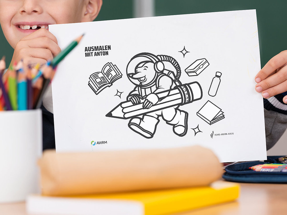 Lächelnder Junge hält im Klassenzimmer ein Ausmalbild mit dem AWRM-Maskottchen Anton. Im Vordergrund stehen bunte Stifte in einem Becher.