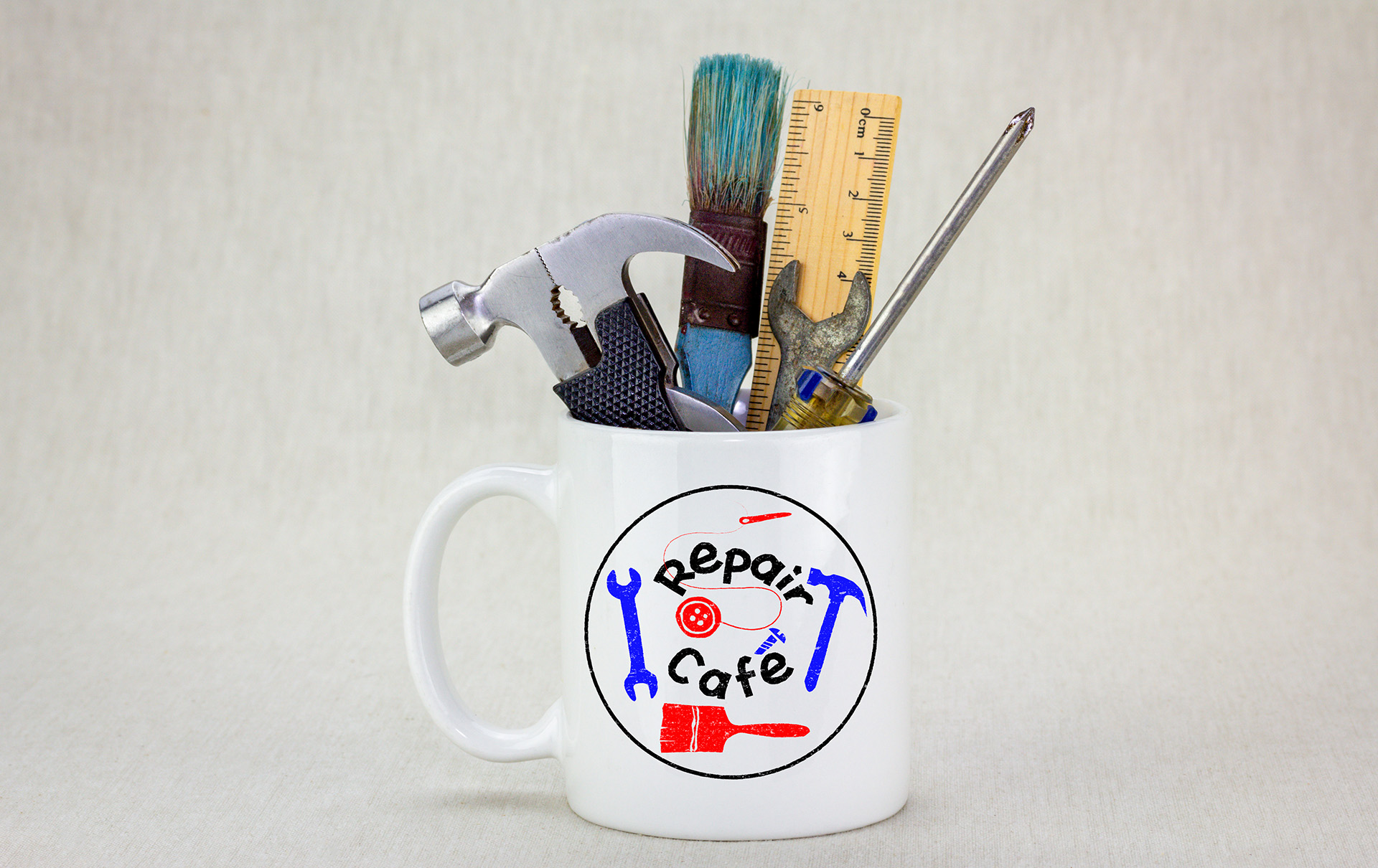 Weiße Tasse mit der Aufschrift „Repair Café“, gefüllt mit Werkzeugen wie Hammer, Schraubenzieher, Pinsel, Lineal und Zange.