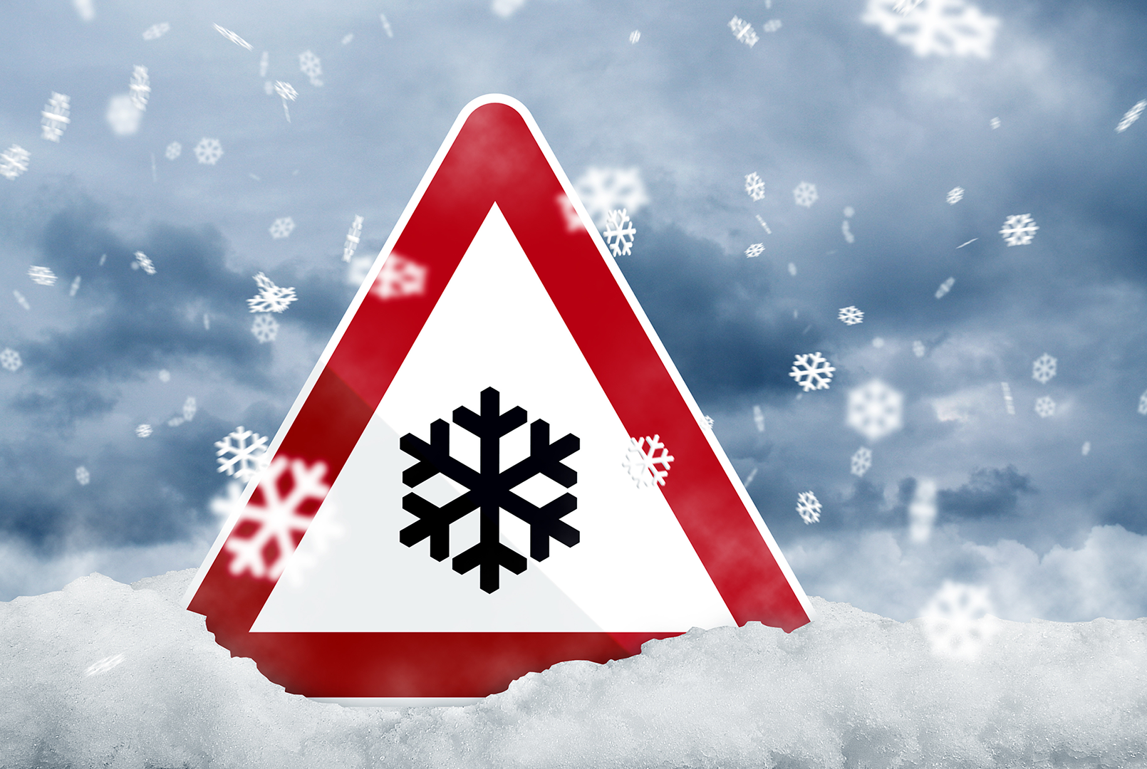 Warnschild mit Schneeflocken-Symbol steckt in einer verschneiten Landschaft, während dichter Schneefall vor einem grau bewölkten Himmel niedergeht.
