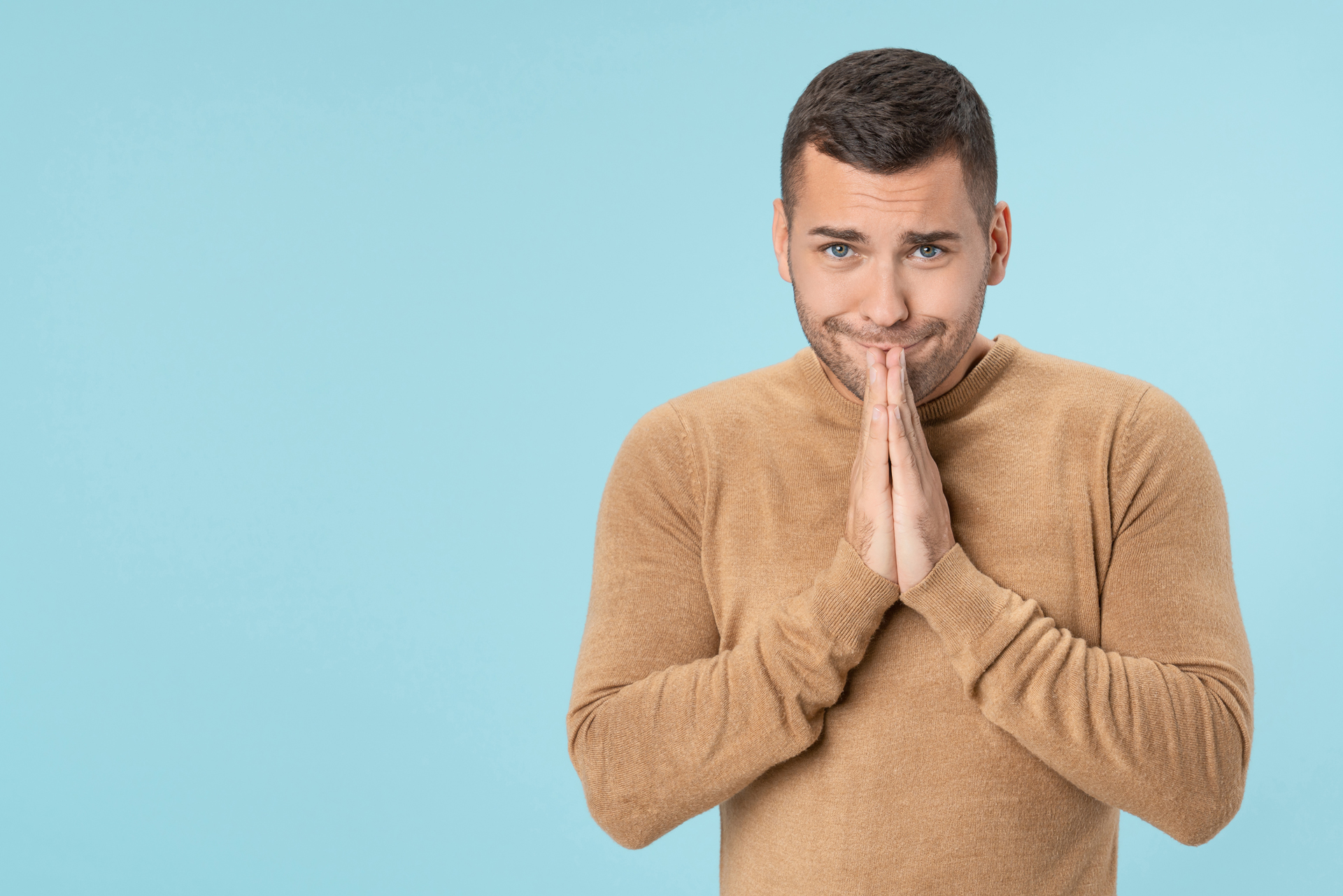 Junger Mann mit braunem Pullover vor hellblauem Hintergrund, der die Hände vor dem Gesicht faltet und entschuldigend schaut.